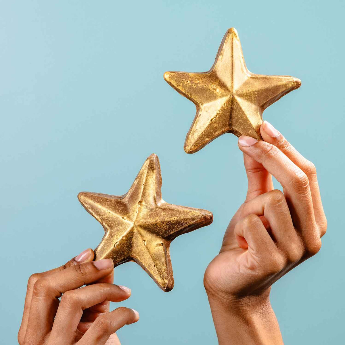 Two hands holding up shimmering gold chocolate stars against a soft blue backdrop – the kind of prize chocolate you actually want to win.