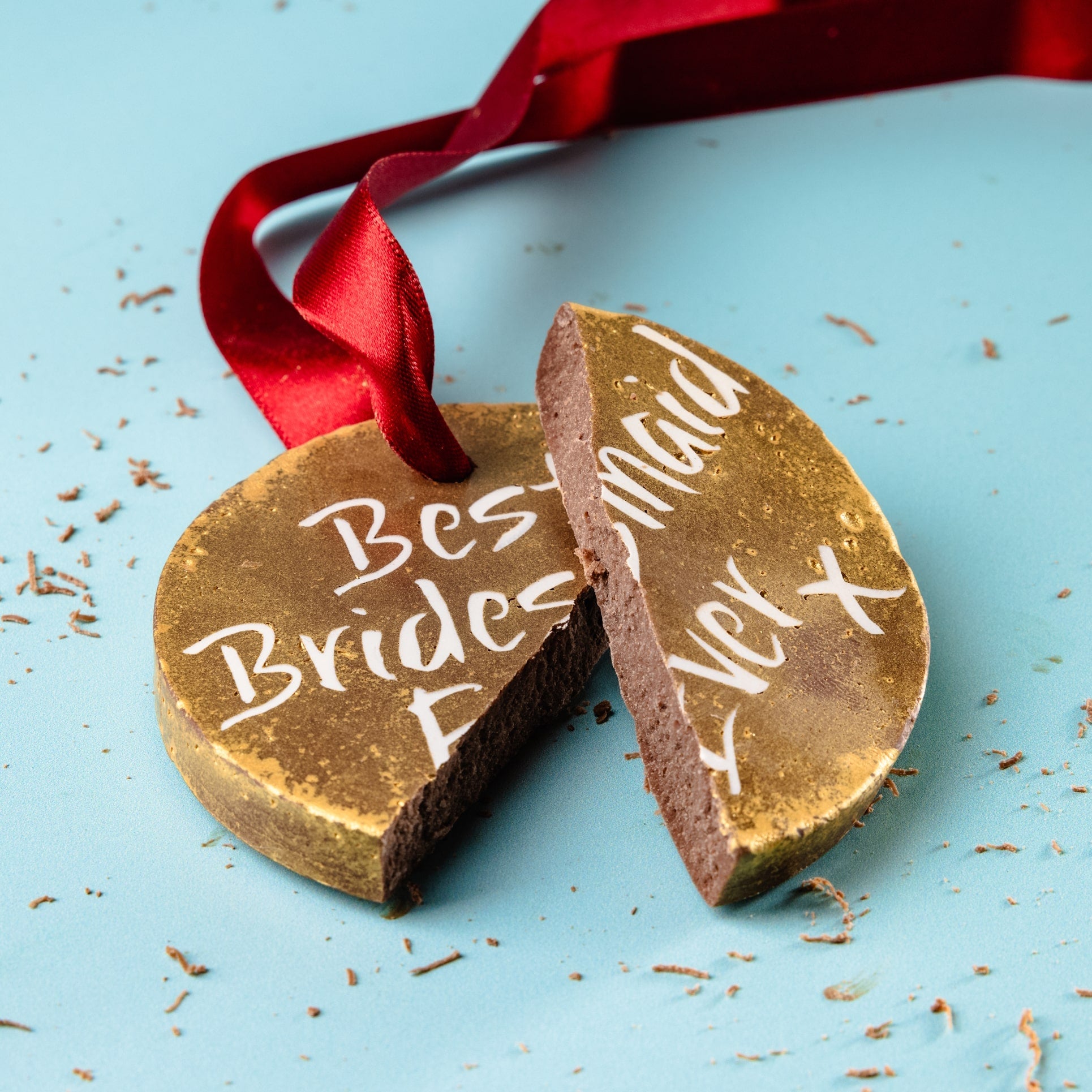 A broken chocolate medal showing the words “Best Bridesmaid Ever”, resting on a blue background with chocolate shavings – a thoughtful and cheeky chocolate gift for bridesmaid duties well done