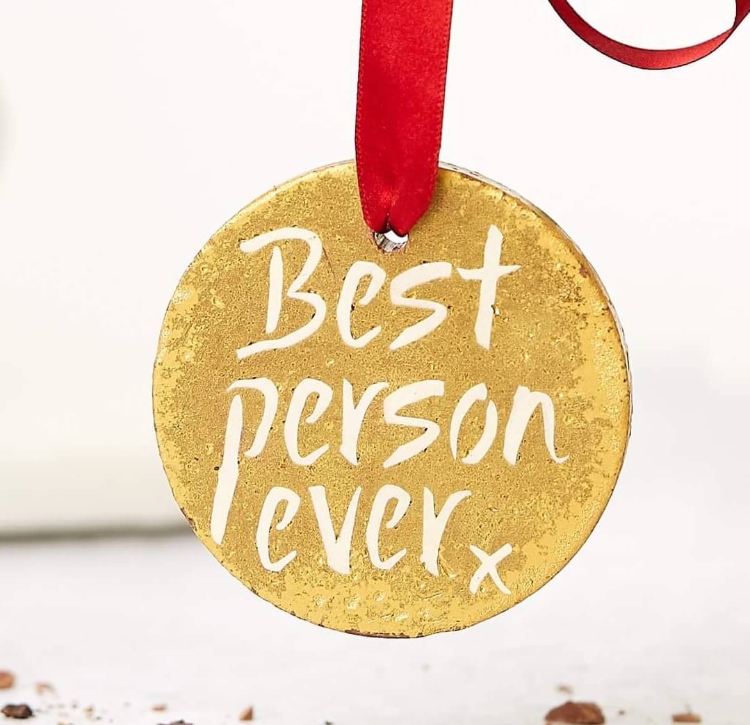 A hand holding a gold medal chocolate with a red ribbon and the words “Best Person Ever” written on it – a sweetly delicious way to say someone’s the greatest.