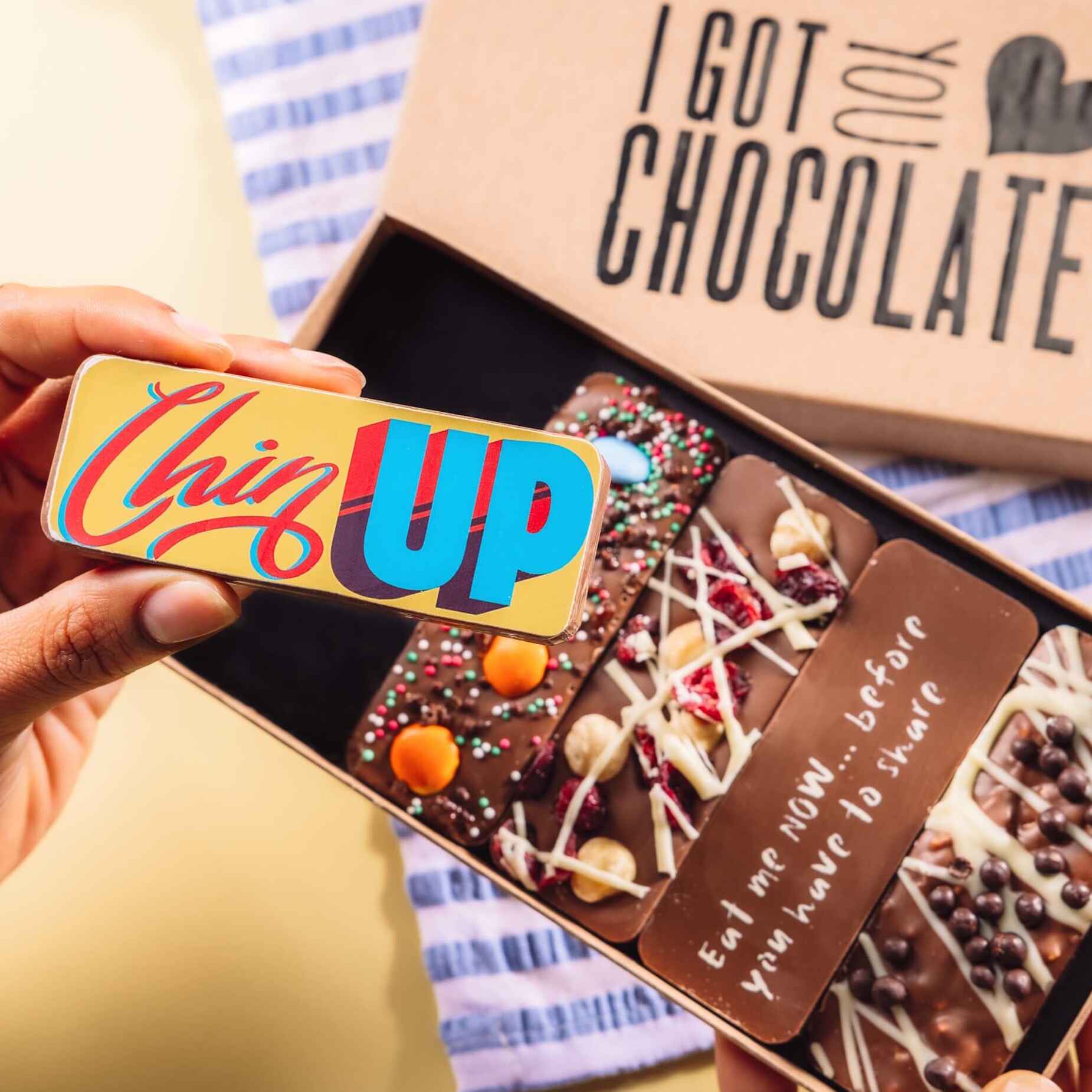 A hand holding a colourful chocolate bar with the phrase "Chin Up" in bold lettering, with Bagstock & Bumble's "I Got You Chocolate" gift box shown in the background. The open box reveals five Belgian chocolate bars with various toppings and message. The perfect chocolate get well gifts.