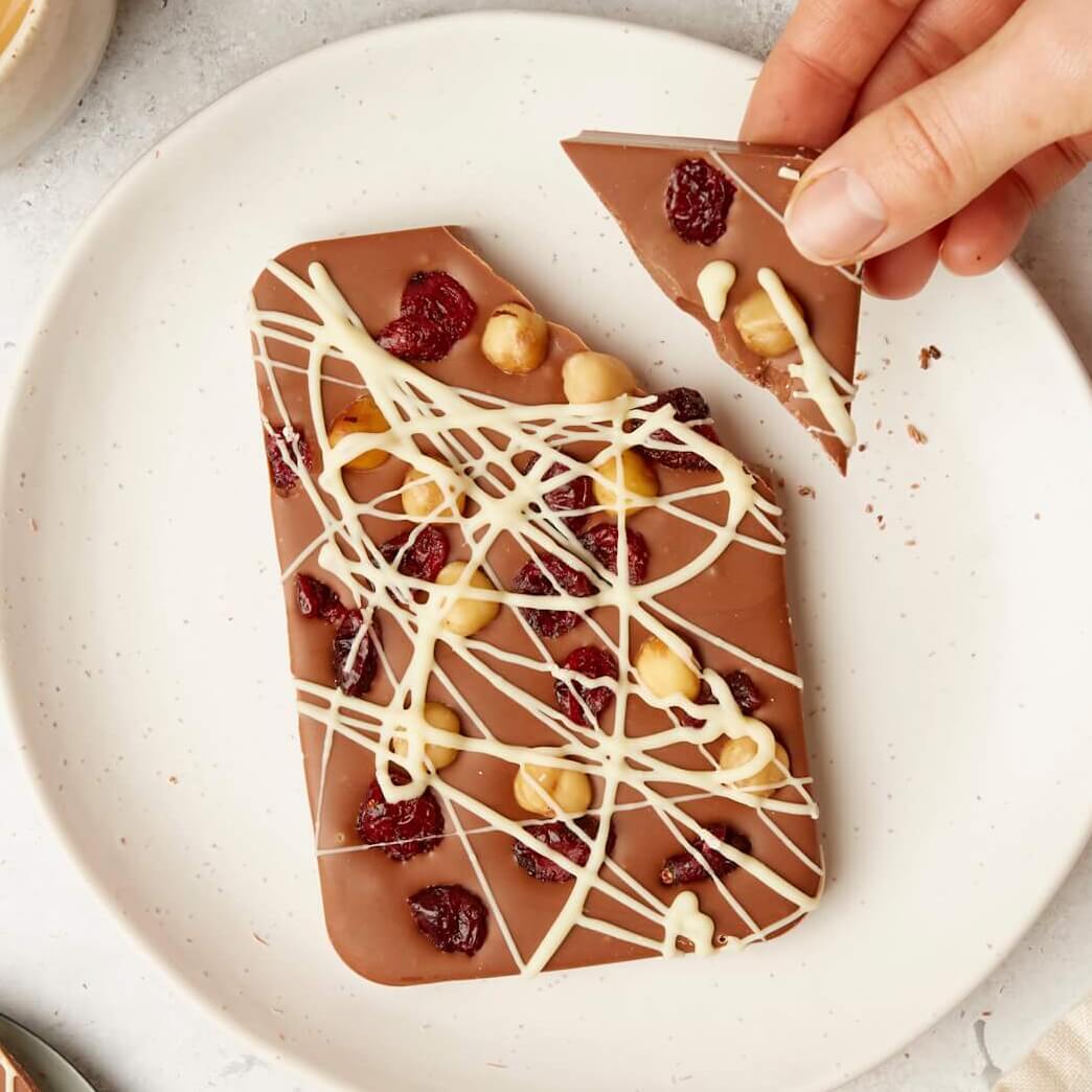 A large fruit and nut chocolate bar topped with whole hazelnuts, dried cranberries, and a drizzle of white chocolate, being broken by hand.