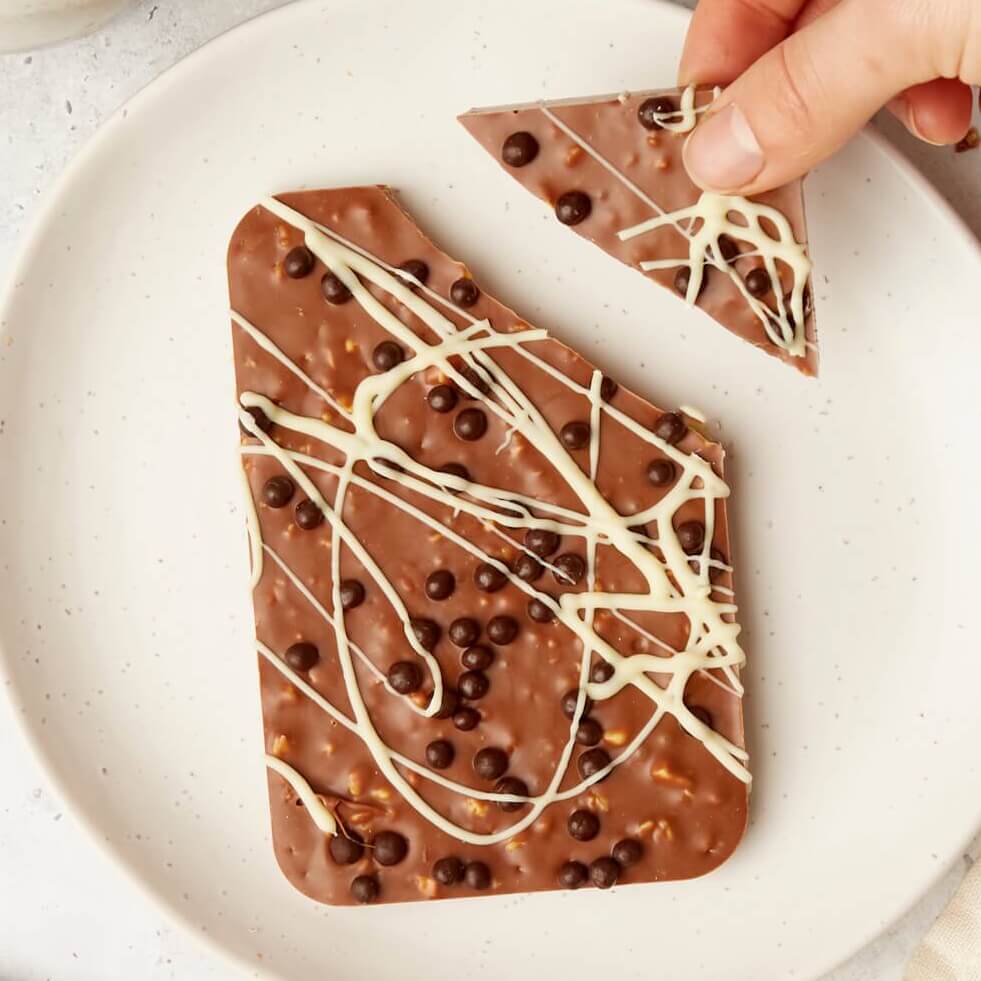 A hand breaks off a triangle from a crunchy milk honeycomb chocolate bar, topped with white chocolate drizzle and crispy chocolate pearls.
