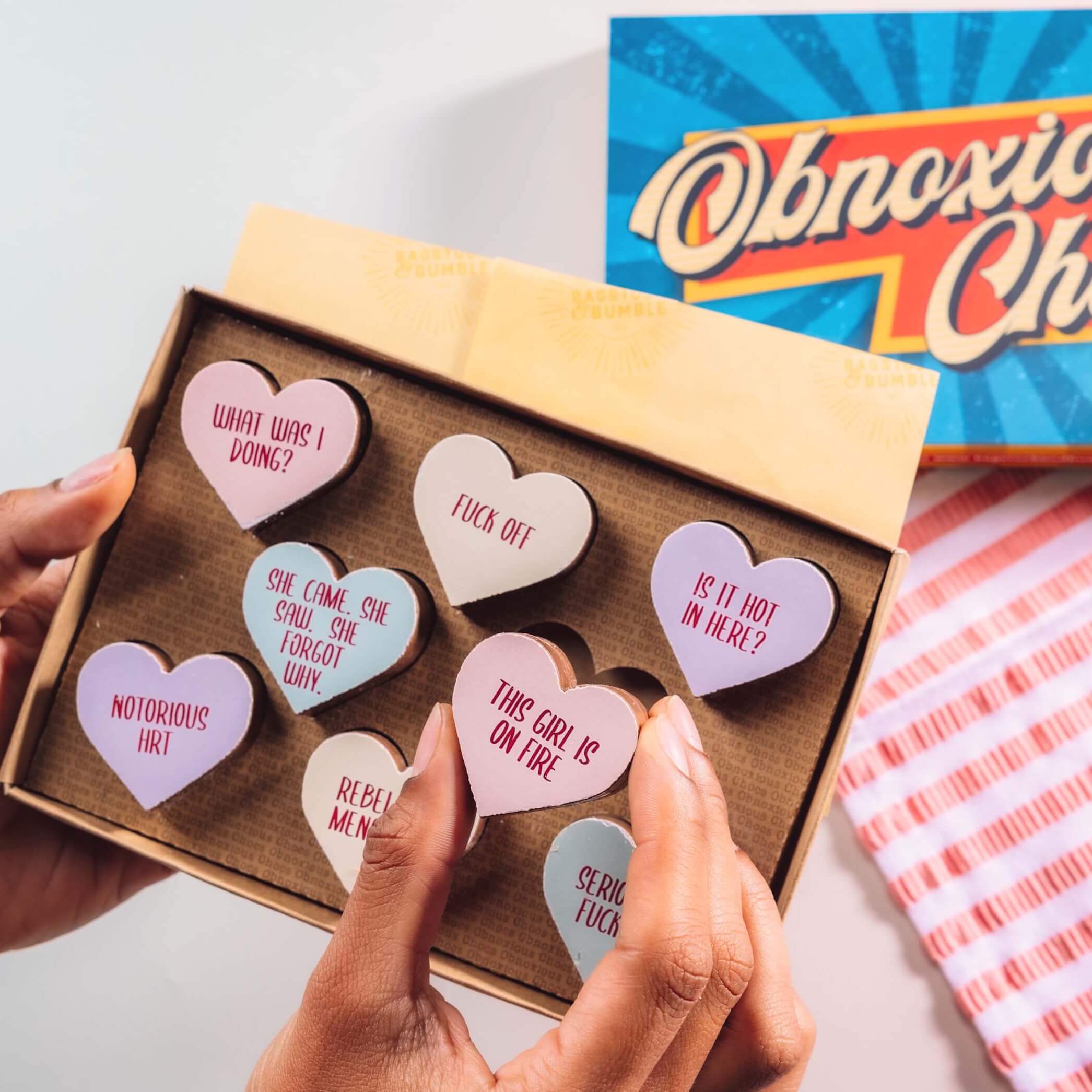 Box of Bagstock & Bumble Menopause Obnoxious Chocs held up, featuring heart-shaped Belgian chocolates with pastel-coloured, sarcastic slogans – a funny menopause gift for friends who need a laugh.