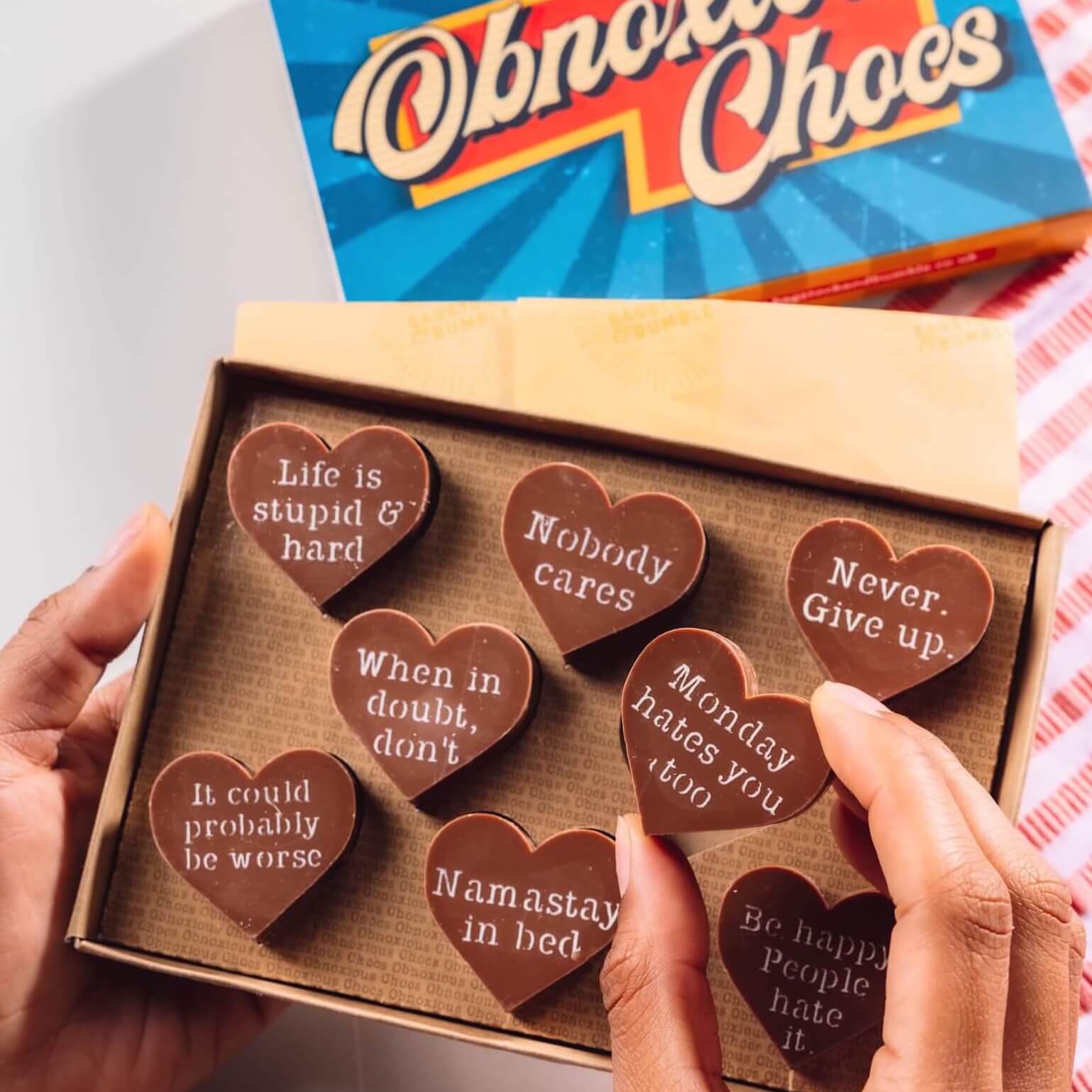 Hand holding a box of Bagstock & Bumble Inspirational Obnoxious Chocs, packed with Belgian chocolate hearts and sarcastic motivational messages.