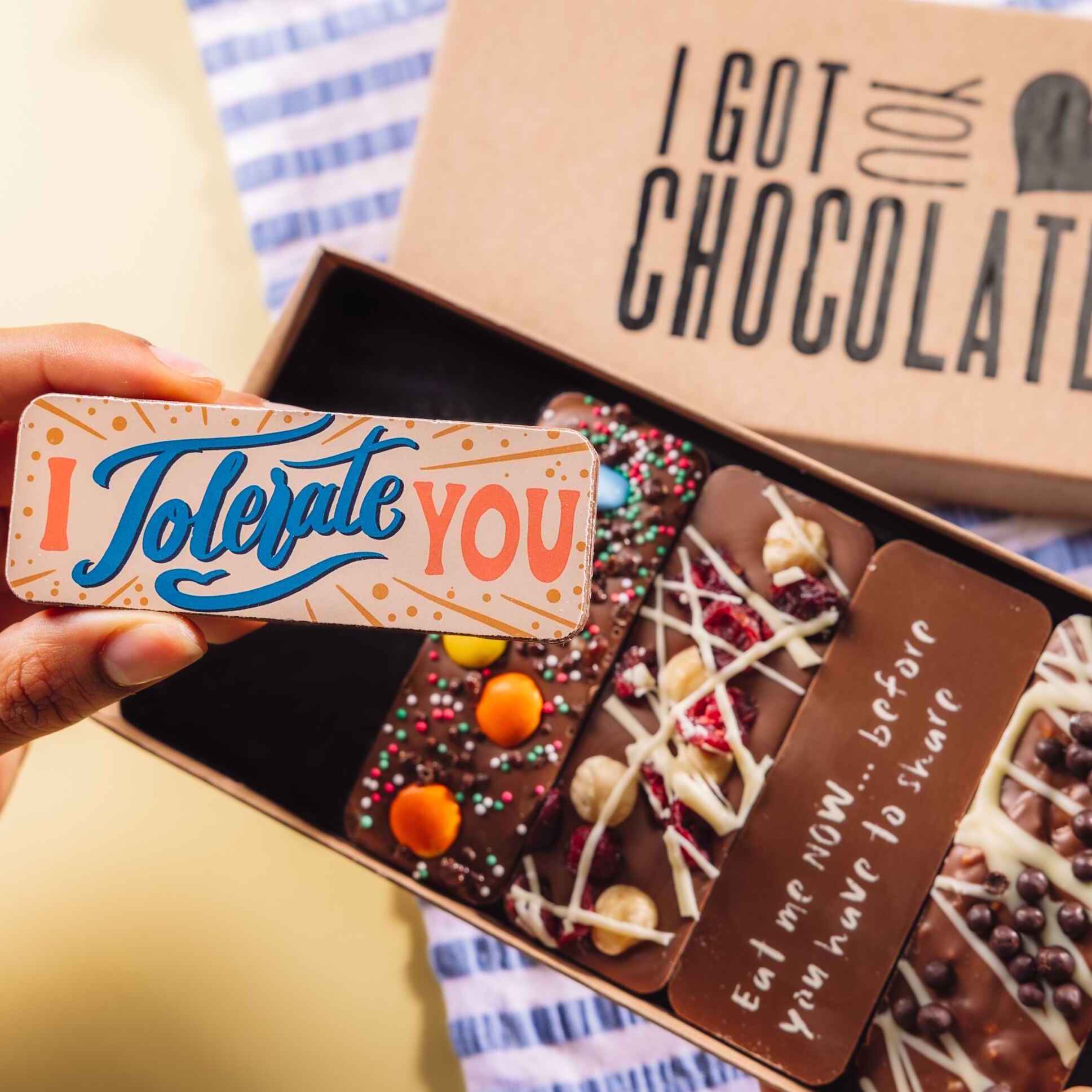 A hand holding a colourful chocolate bar with the message “I Tolerate You” above an open gift box of other bars – a perfect pick for funny couples gifts.
