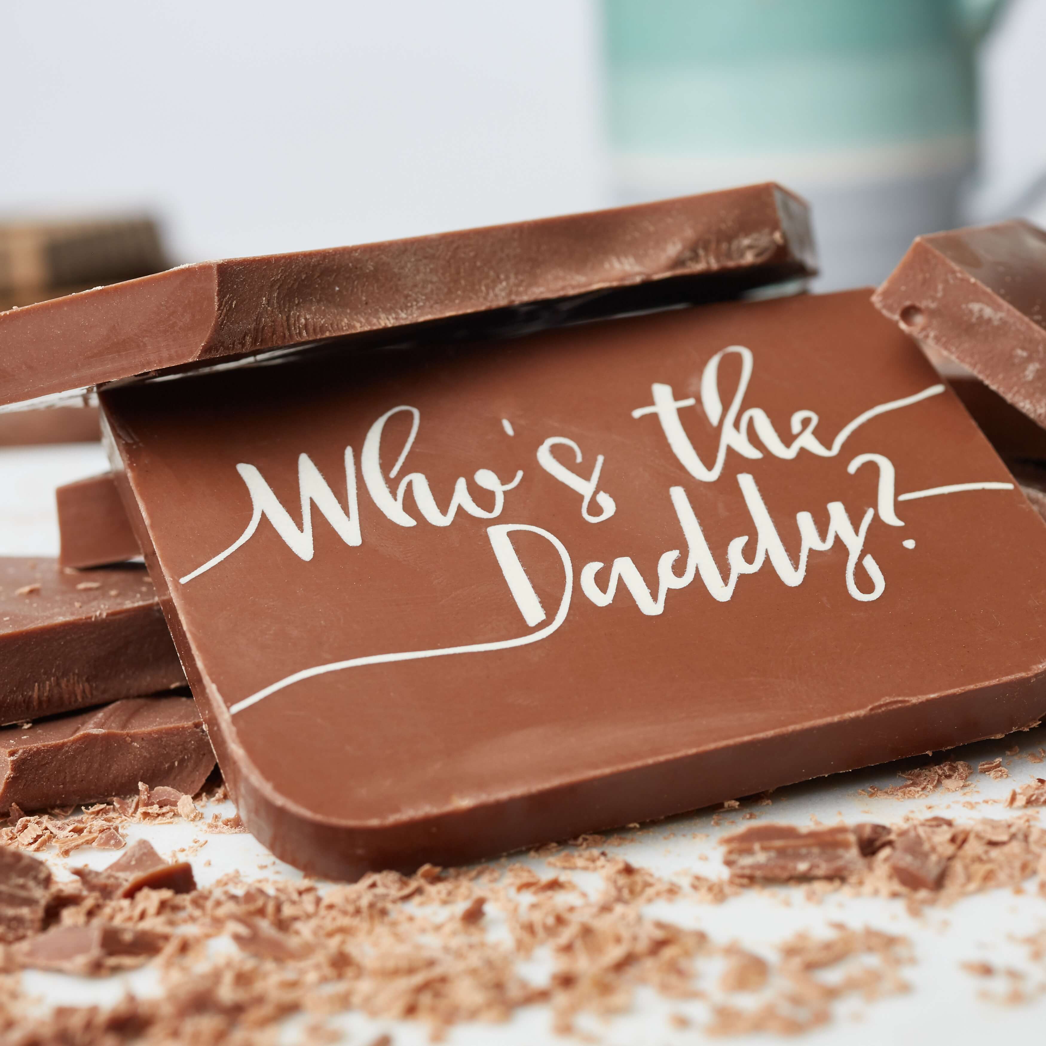 Close-up of a chunky milk chocolate bar with ‘Who’s the Daddy?’ written in white cocoa butter, surrounded by chocolate pieces. The perfect Father's Day Chocolate GIft.