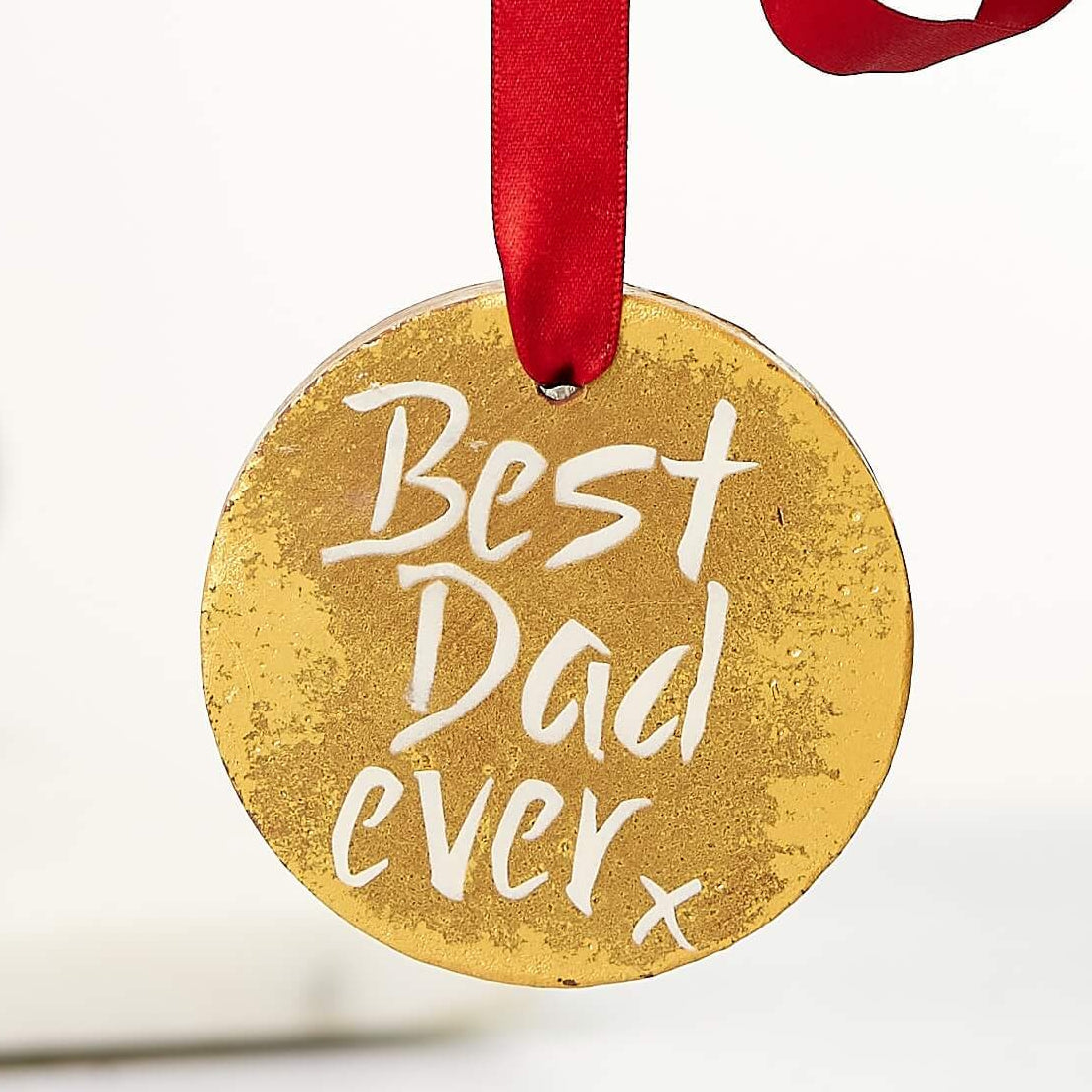 A hand holding up a gold chocolate medal with a red ribbon and the words “Best Dad Ever” written on it — the perfect edible medal for best dad, no speech required.