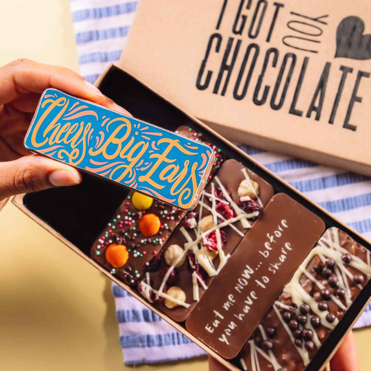 Hand holding a colourful chocolate bar with the message “Cheers Big Ears”, in front of an open gift box of handmade chocolate bars – a fun and thoughtful option for chocolate thank you gifts.