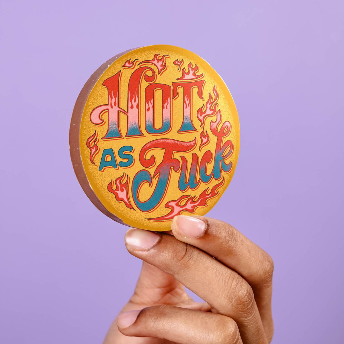 Cheeky, funny valentines chocolate disc made from milk chocolate with a colourful edible transfer reading “Hot As Fuck”, held in hand against a lilac background.