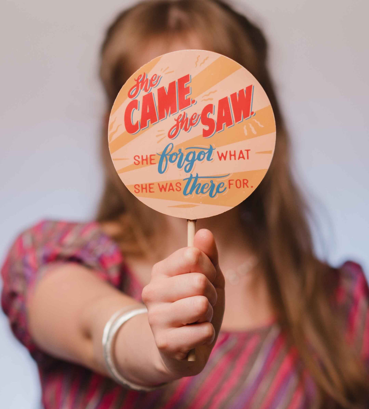 An out of focus body holding a jumbo lolly up to camera.  The lolly has a colourful chocolate transfer with a colourful, retro styled ‘She came, she saw, she forgot what she was there for’ slogan.