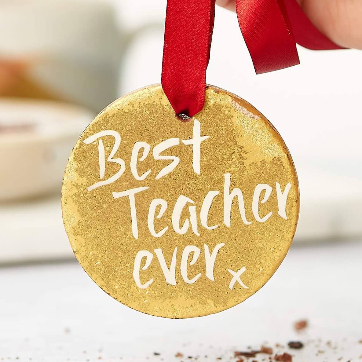 A hand holding a gold chocolate medal with a red ribbon and the words “Best Teacher Ever” written on it – a sweet and funny option for the best teacher gift going.