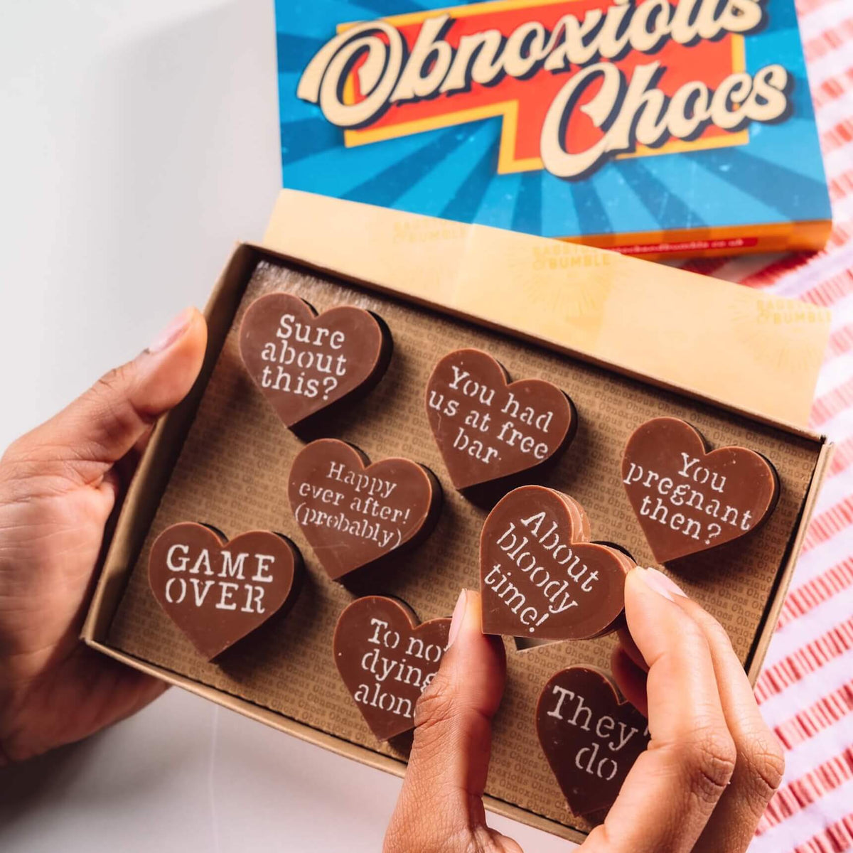 Bagstock & Bumble Engagement Obnoxious Chocs gift box being held up, filled with Fairtrade chocolate hearts decorated with sarcastic messages for the soon to be nearly-weds.