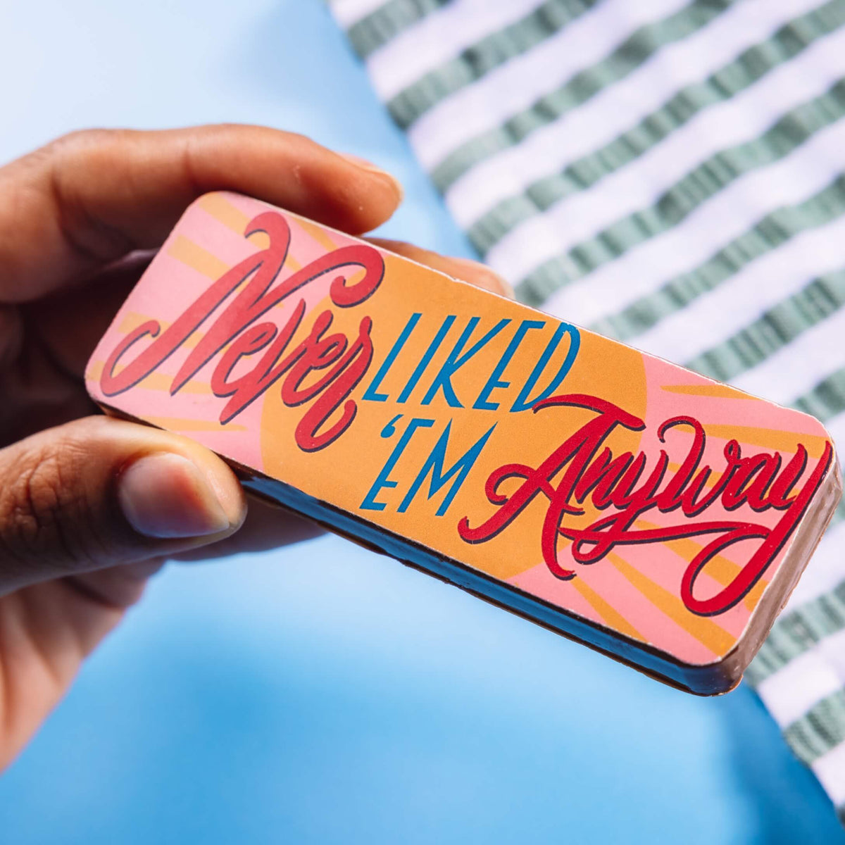 Close-up of a hand holding a chocolate bar that reads “Never Liked ’Em Anyway” in bold, colourful script – add it to a breakup care package to put a smile on their face.
