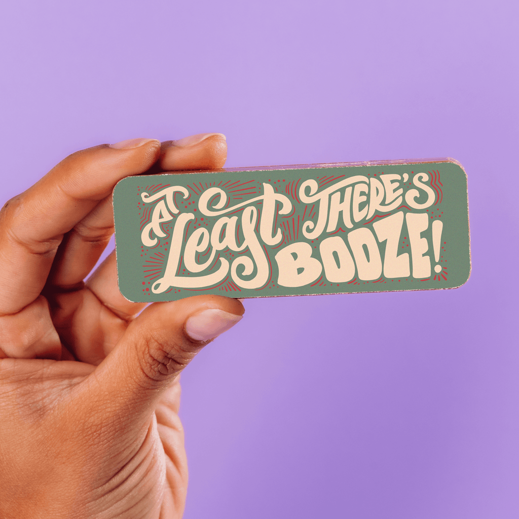 Hand holding funny Christmas chocolate bar that reads "At least there's booze" in beige and red font on a green background.