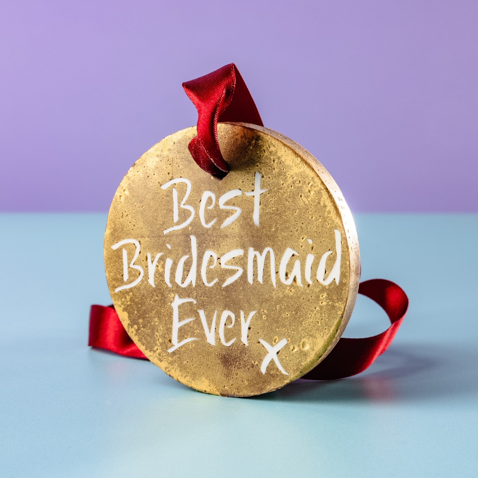 A gold chocolate medal with red ribbon reading “Best Bridesmaid Ever”, propped up on a pastel background – perfect for quirky and delicious bridesmaid favours.
