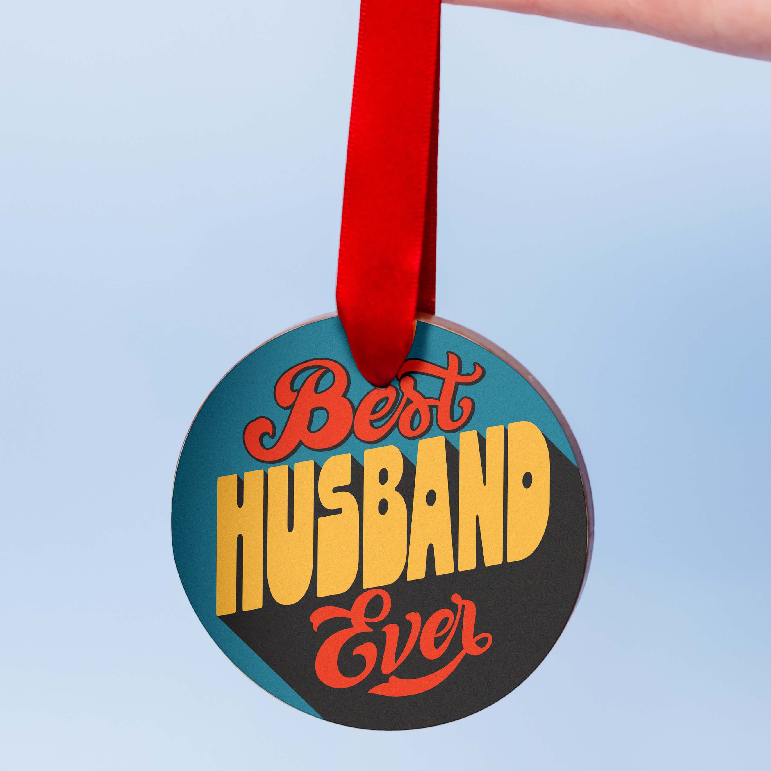 Chocolate valentines gift for husband, a chunky milk chocolate medal with a colourful edible transfer reading “Best Husband Ever”, hanging from a red ribbon against a pale blue background.