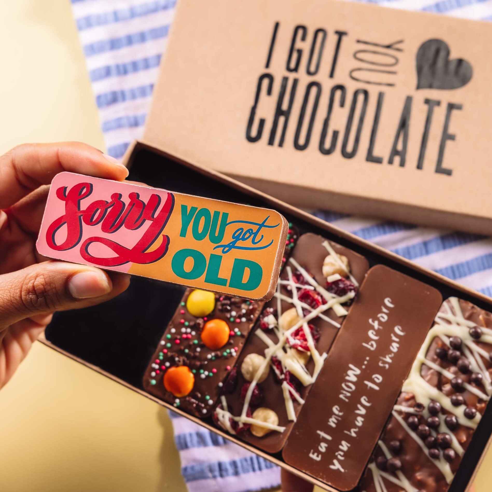 Hand holding a colourful chocolate bar with the message “Sorry You Got Old”, in front of an open birthday chocolate box.  The box features five handmade chocolate bars with cheeky messages and yummy toppings.