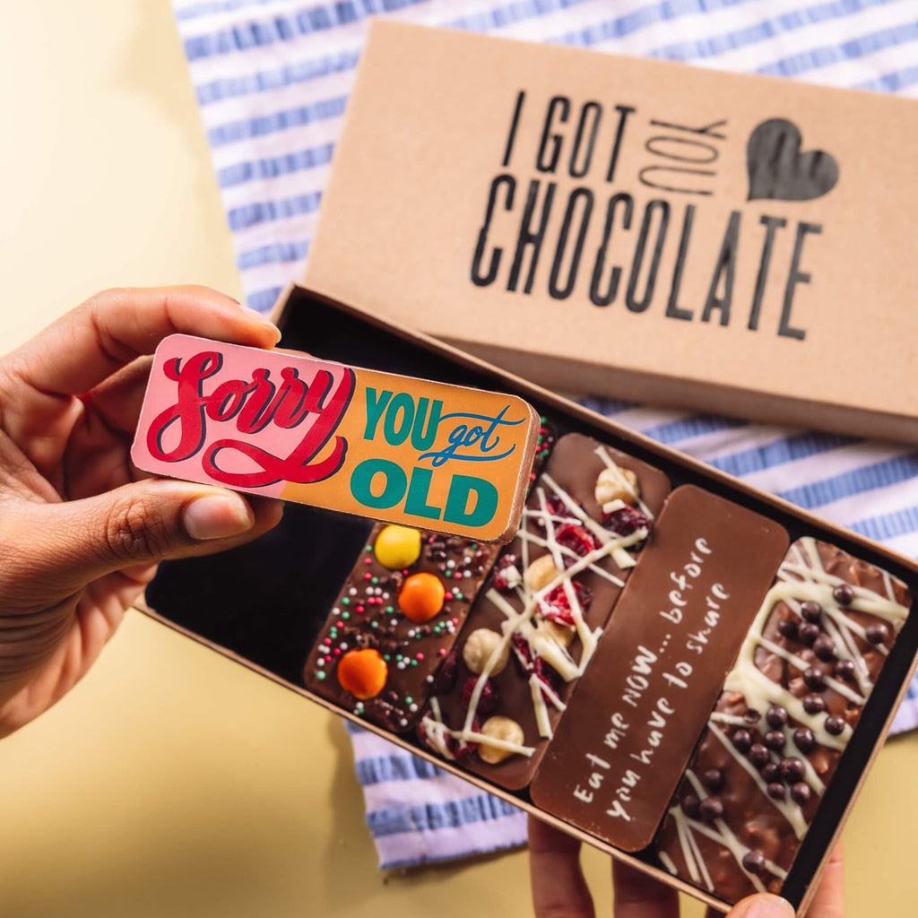 Hand holding a colourful chocolate bar with the message “Sorry You Got Old”, in front of an open birthday chocolate box.  The box features five handmade chocolate bars with cheeky messages and yummy toppings.