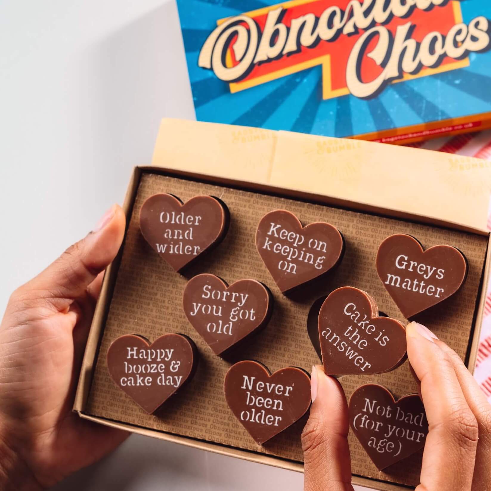 Gift box of Bagstock & Bumble Happy Birthday Obnoxious Chocs held up, featuring heart-shaped Fairtrade chocolate with sarcastic birthday phrases, ideal for anyone getting older (and grumpier).