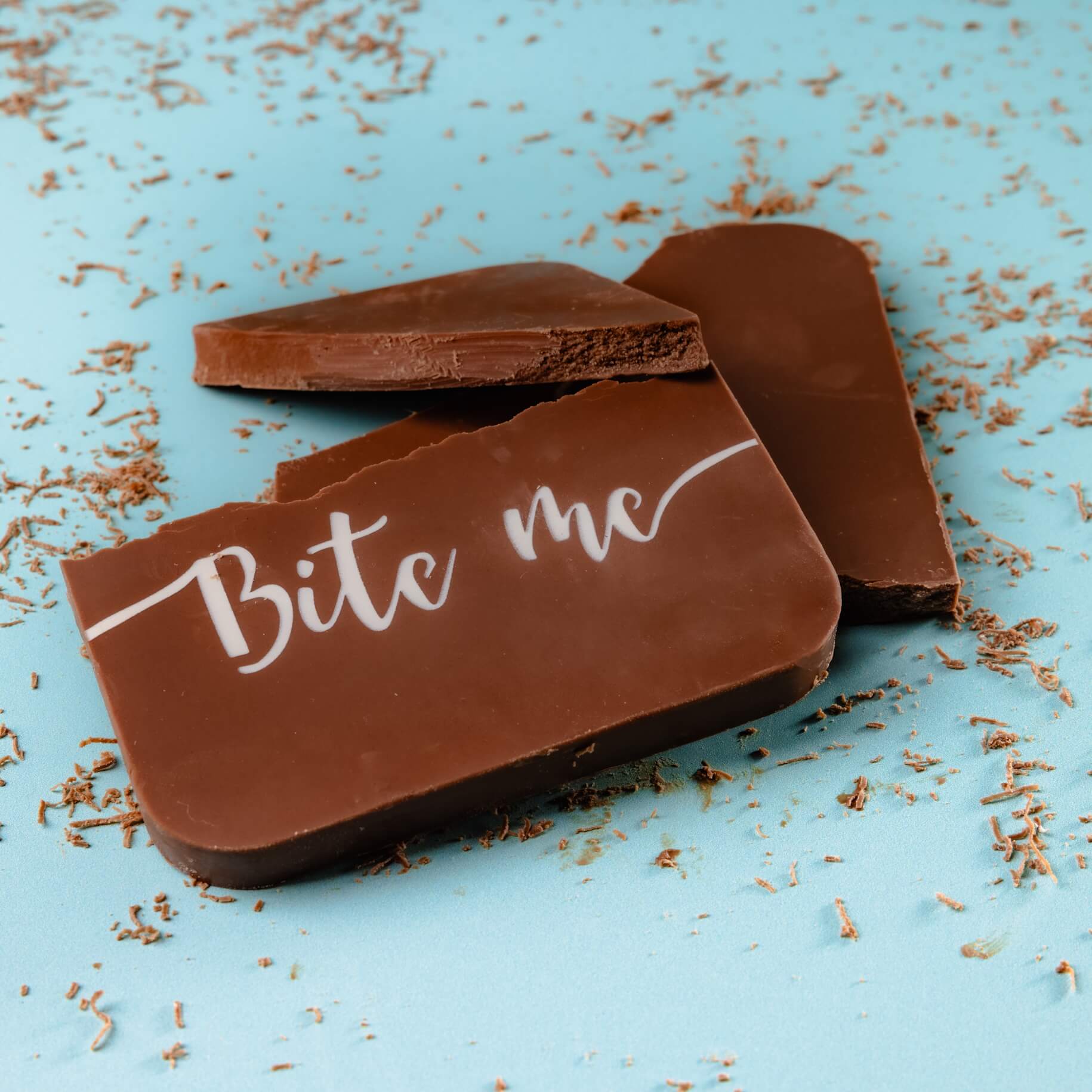 Chocolate to Your Door - Close-up of a snapped “Bite me” chocolate bar on a blue background, surrounded by chocolate shavings.  Deliciously passive aggressive.