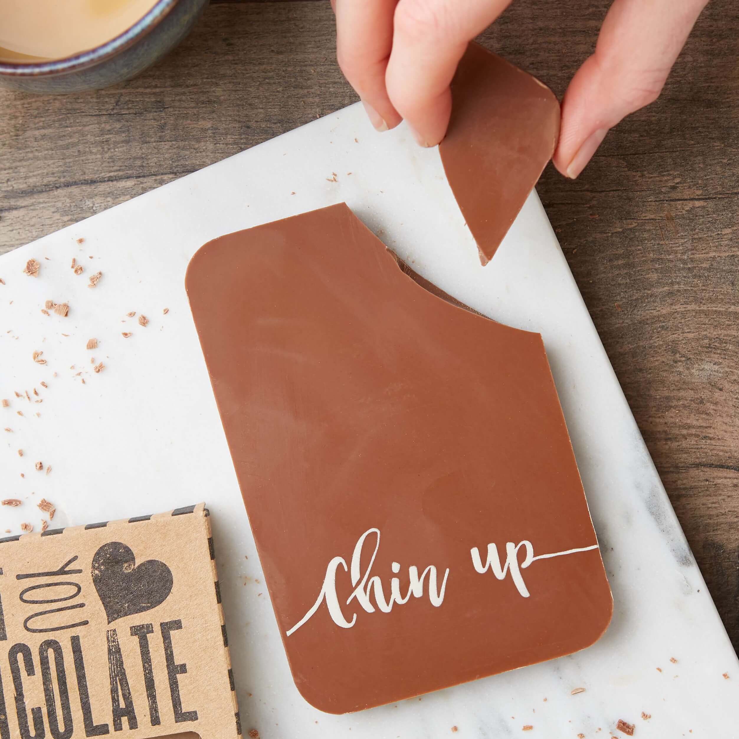 A hand breaking a piece off a “Chin up” milk chocolate bar next to its empty packaging – the ultimate cheer up chocolate gift 