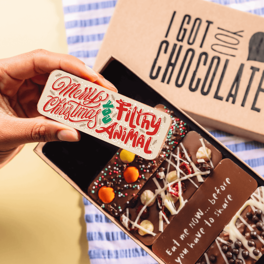 Close-up of a hand holding a "Merry Christmas, you filthy animal" bar above an open Christmas chocolate selection box containing several custom-made, decorated chocolate bars.