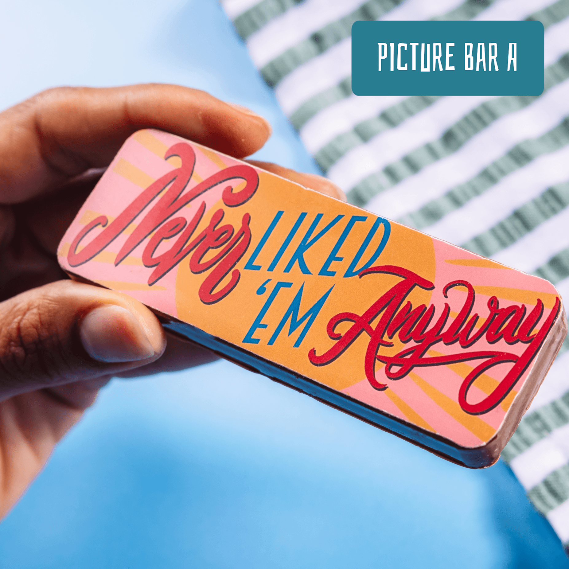 Close-up of a hand holding a chocolate bar that reads “Never Liked ’Em Anyway” in bold, colourful script – add it to a breakup care package to put a smile on their face.