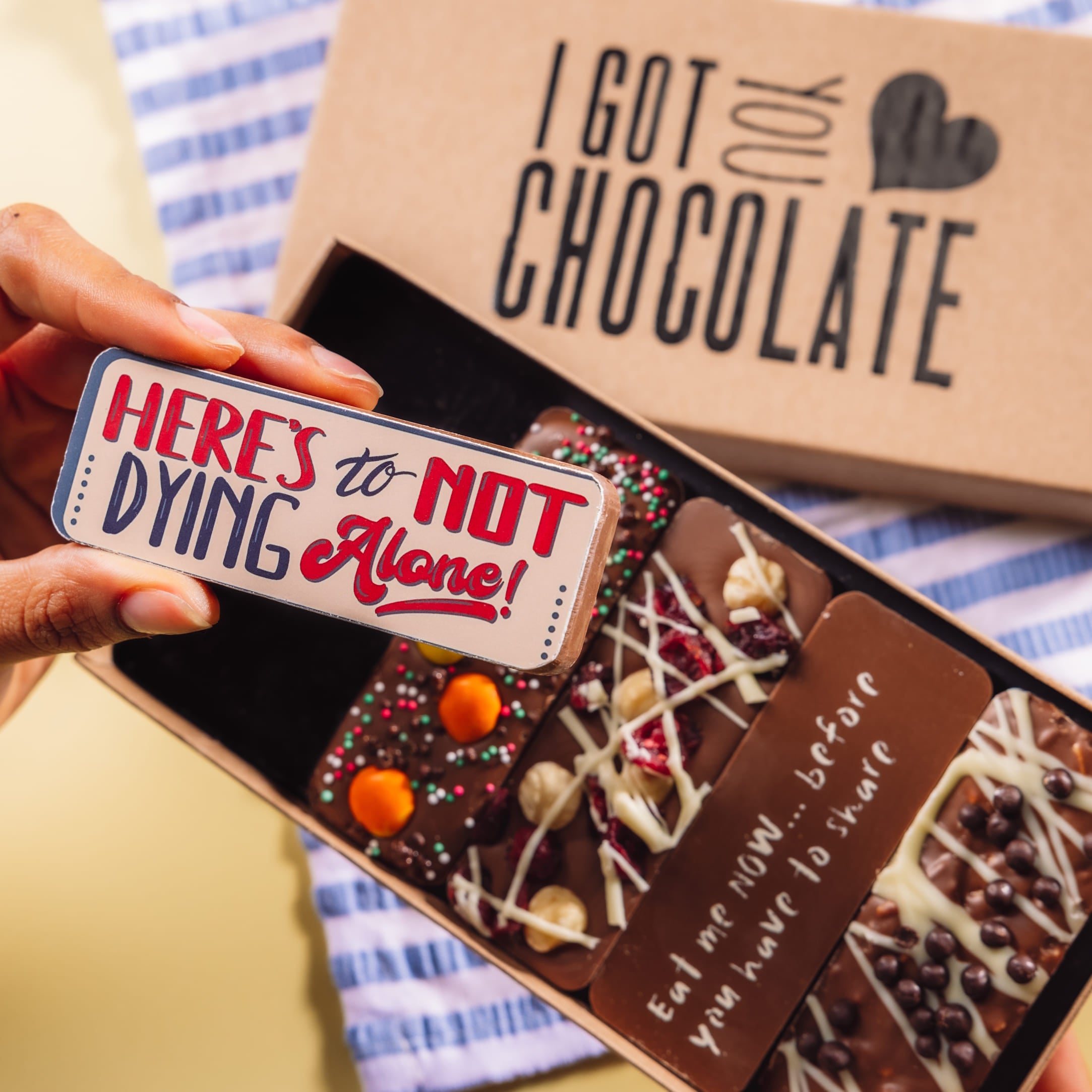 A hand holding a chocolate bar that reads “Here’s to not dying alone!” from Bagstock & Bumble’s “I Got You Chocolate” gift box. The perfect engagement chocolates for couples with a sense of humour.