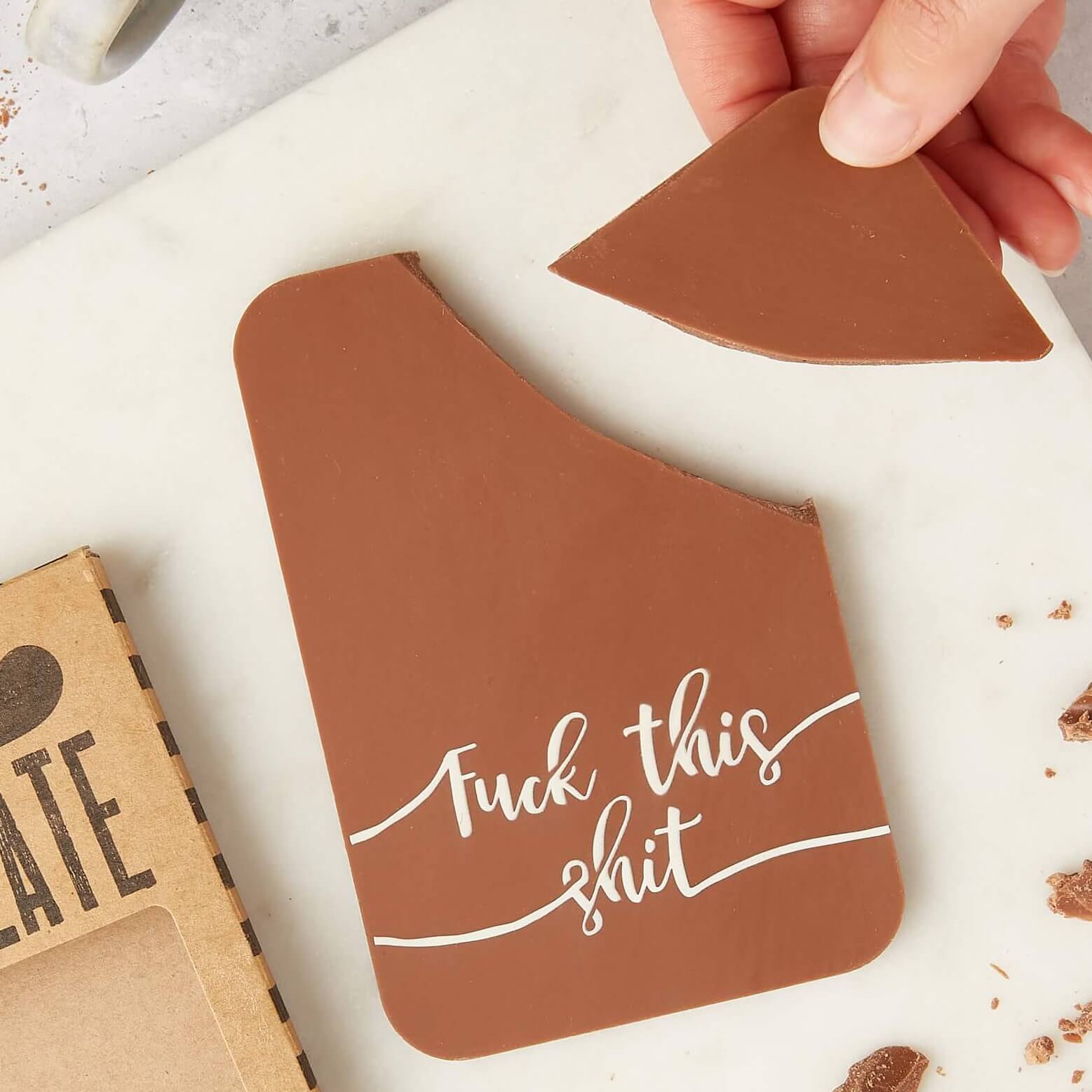 A hand holding a broken piece of a milk chocolate bar with “Fuck this shit” written on it – one of our funny chocolate bars for when words aren’t quite enough.