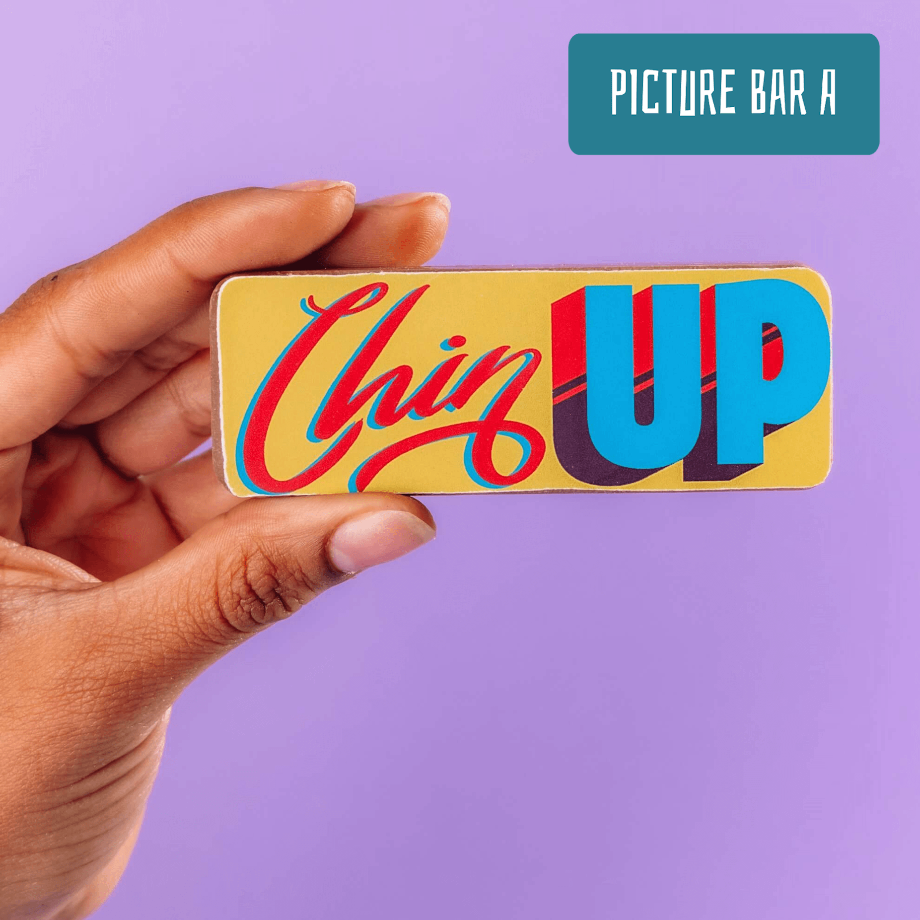 Close-up of a hand holding a vibrant chocolate bar that reads "Chin Up" in playful, retro-style typography. A light-hearted and cheeky addition to Bagstock & Bumble’s funny get well soon gifts range.
