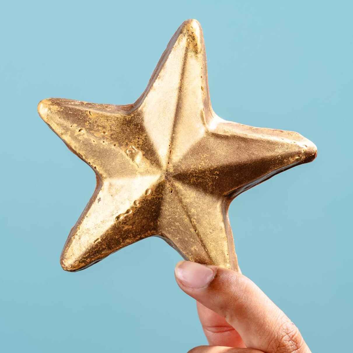 A hand holding a shimmering chocolate gold star against a blue background – a shiny, edible pat on the back for someone who’s nailed it.