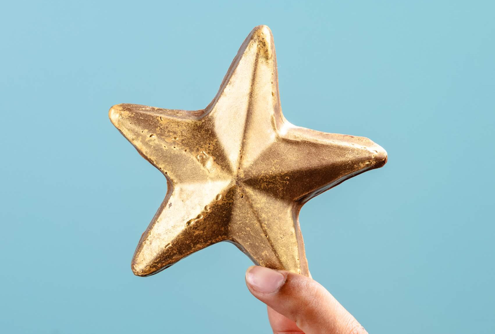 A hand holding a shimmering chocolate gold star against a blue background – a shiny, edible pat on the back for someone who’s nailed it.
