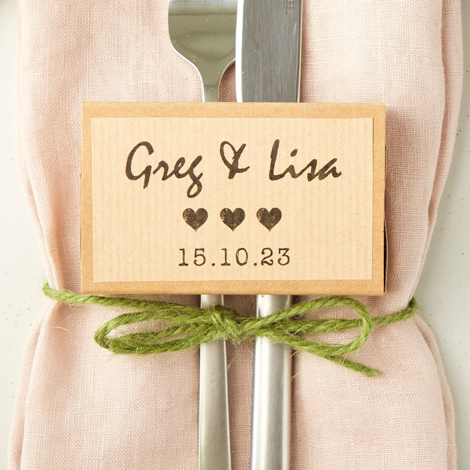 Gift box personalised with 'Greg & Lisa' plus a date, pictured on a light pink napkin and knife and fork.