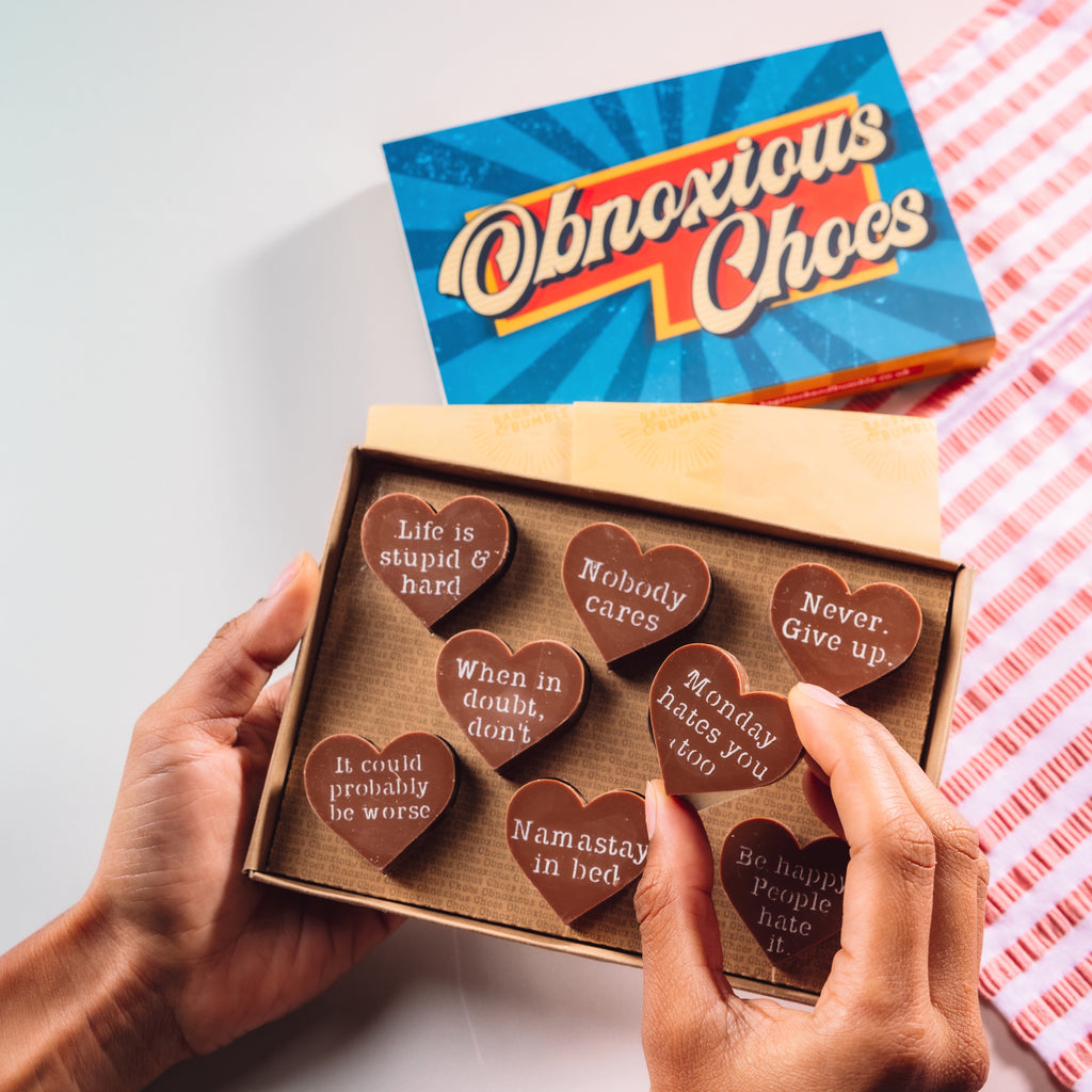 Hand holding a box of Bagstock & Bumble Inspirational Obnoxious Chocs, packed with Belgian chocolate hearts and sarcastic motivational messages.