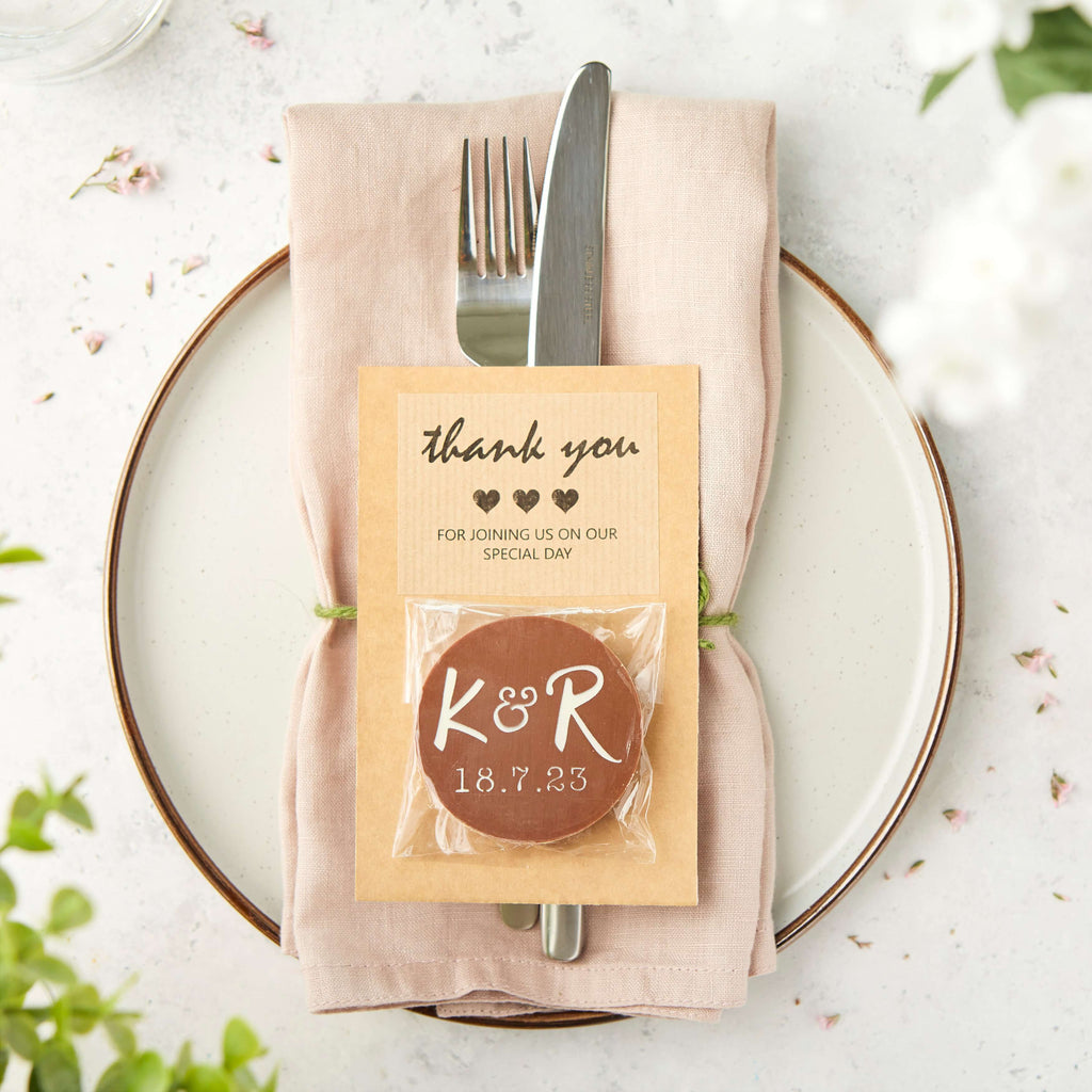 Place setting with folded napkin, cutlery, and a custom chocolate with K&R initials attached to a 'thank you' card.