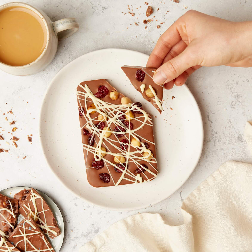 A large fruit and nut chocolate bar topped with whole hazelnuts, dried cranberries, and a drizzle of white chocolate, being broken by hand.