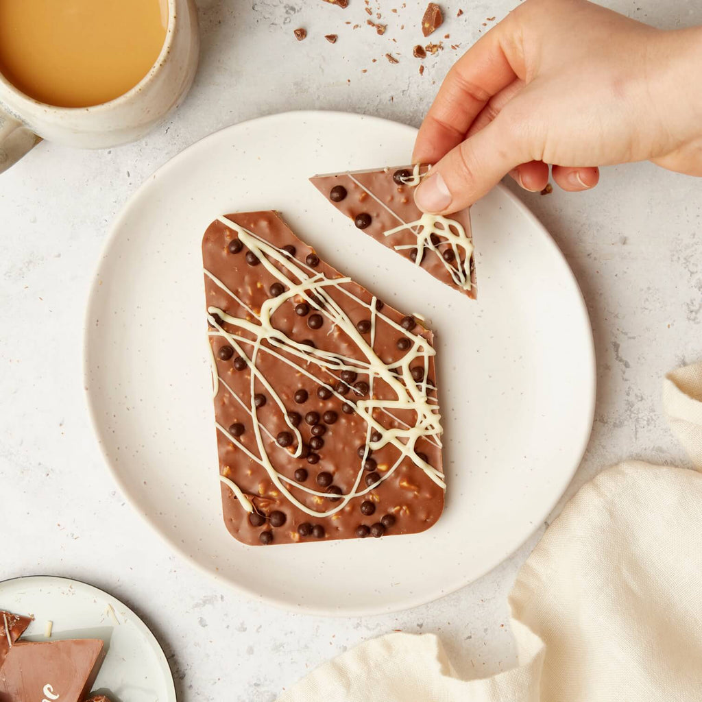 A hand breaks off a triangle from a crunchy milk honeycomb chocolate bar, topped with white chocolate drizzle and crispy chocolate pearls.