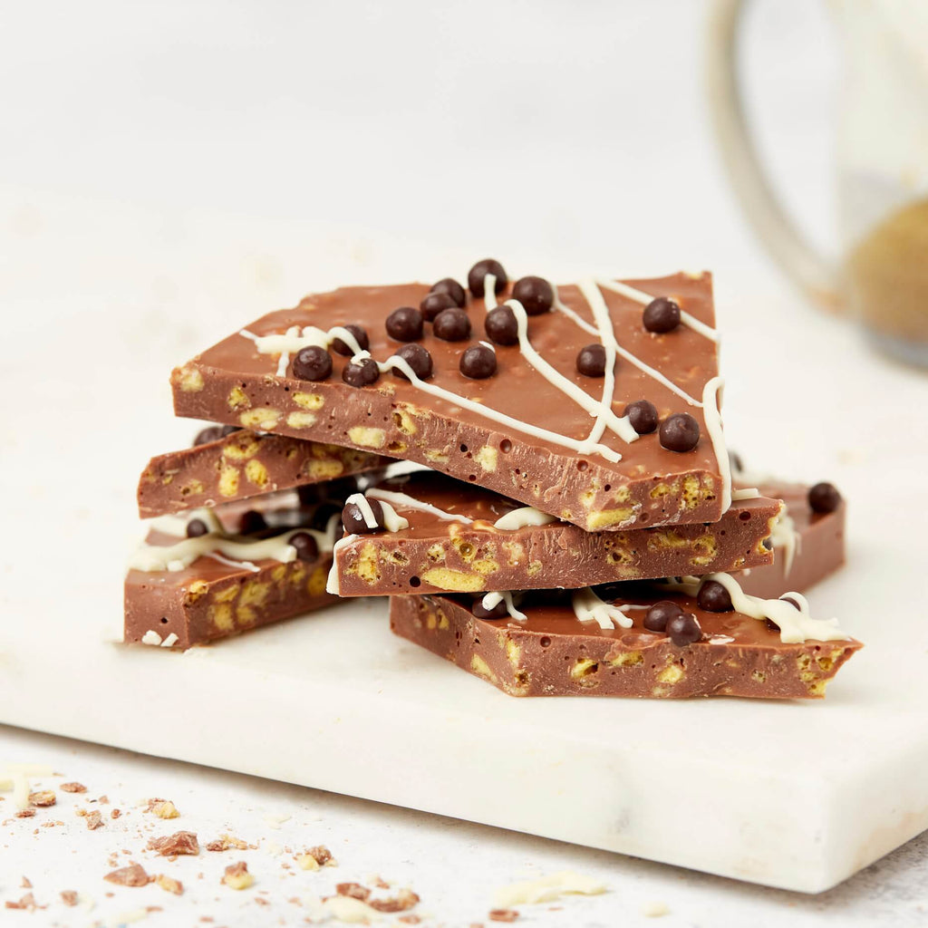 A stack of handmade milk chocolate pieces filled with honeycomb and topped with dark chocolate crispearls and white drizzle – a delicious bite of artisan chocolate UK style.