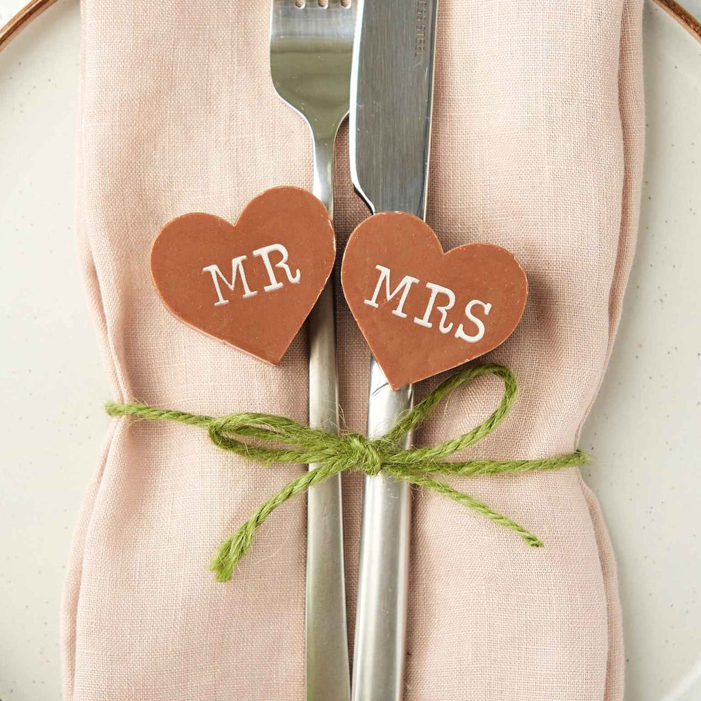 Place setting with 'MR' and 'MRS' heart-shaped custom chocolates on a knife and fork and a pink napkin.