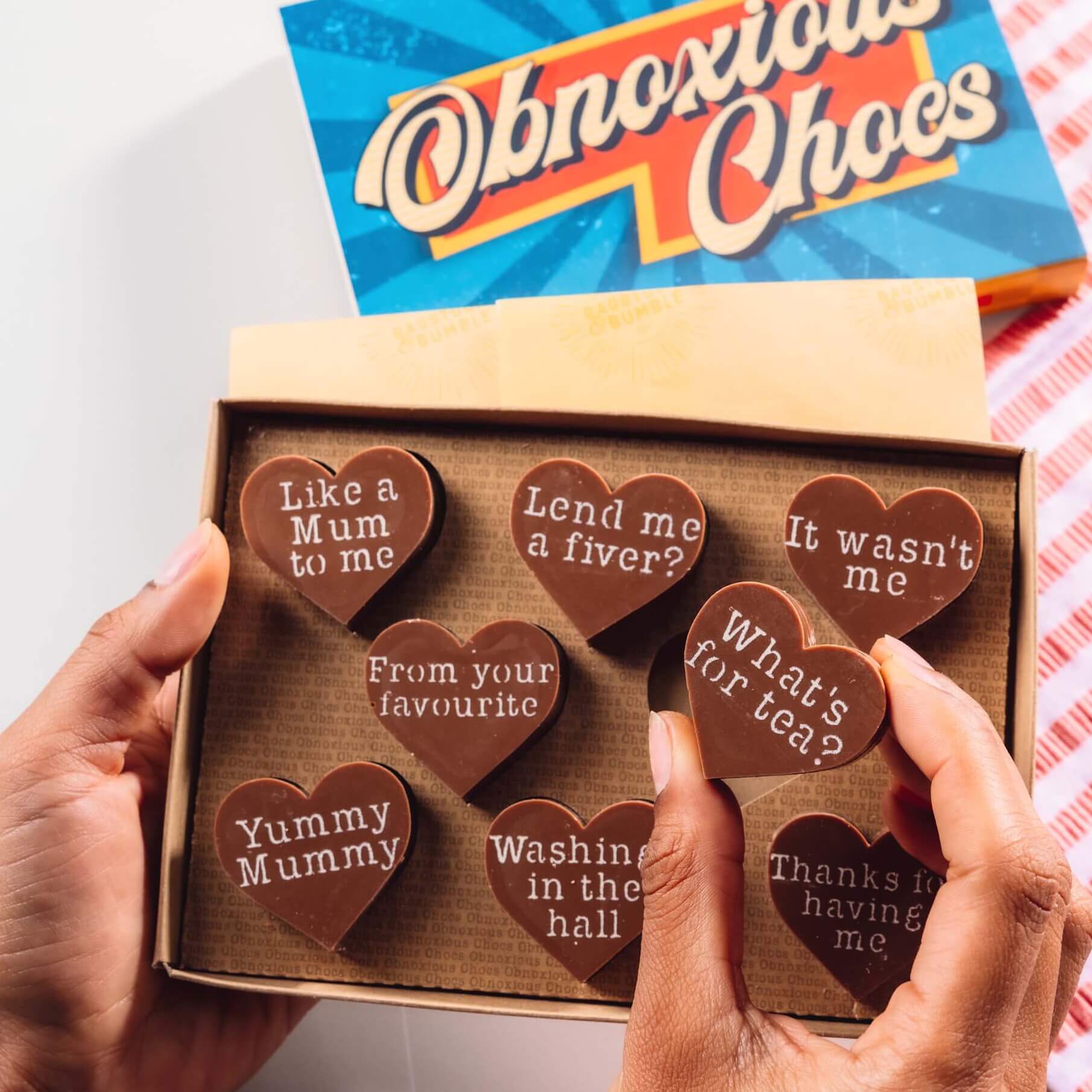Hands holding a box of Bagstock & Bumble Mum Obnoxious Chocs, packed with heart-shaped Fairtrade chocolates featuring witty, no-nonsense messages for mums everywhere.