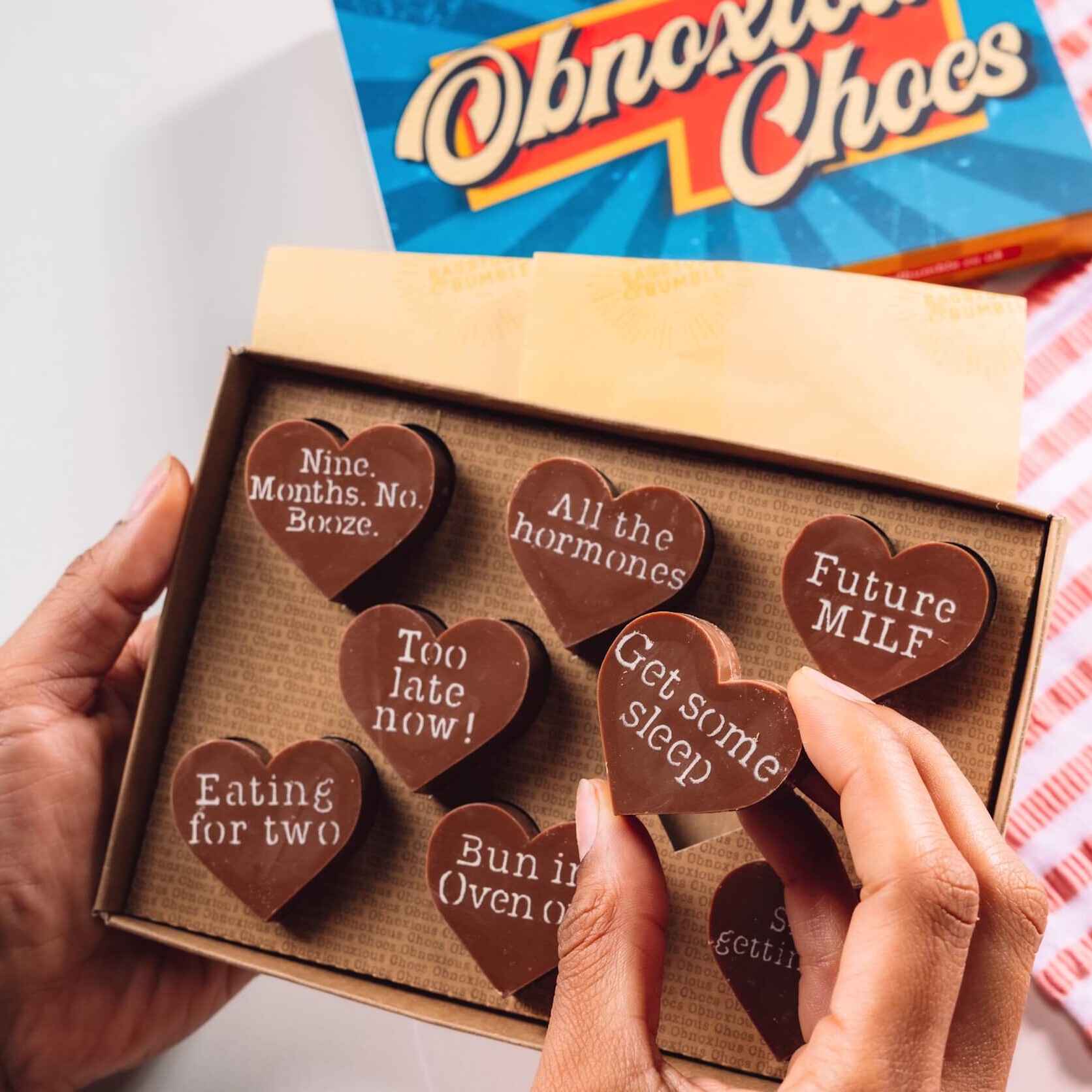 Bagstock & Bumble Obnoxious Chocs gift box featuring heart-shaped Fairtrade Belgian chocolates engraved with cheeky pregnancy sayings like 'Future MILF' and 'Get some sleep', a fun and unusual present for expectant mums.