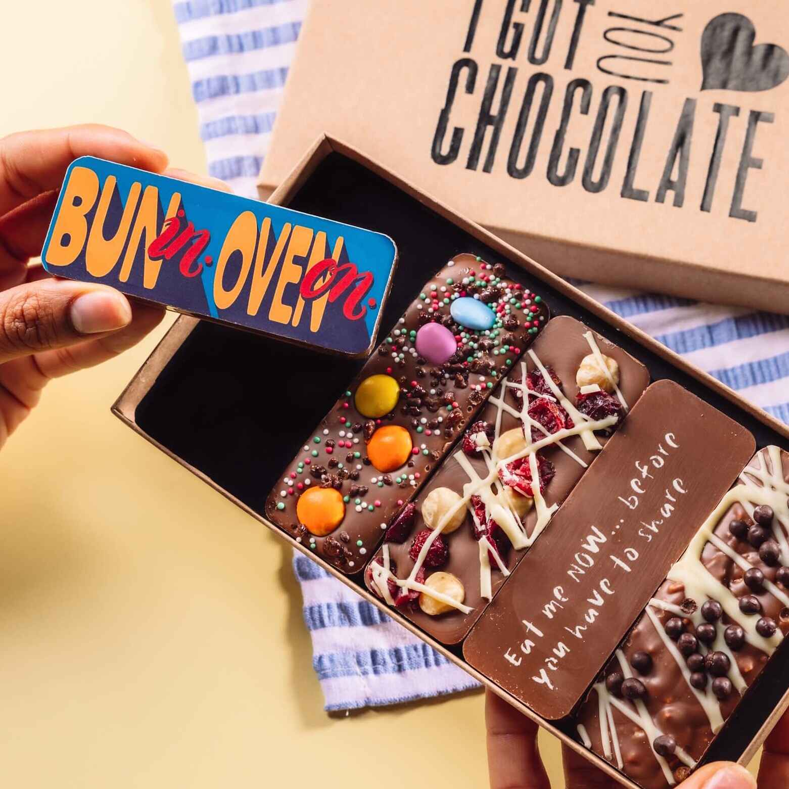A hand holding a mini chocolate bar that reads “Bun in. Oven on.” above an open box of assorted chocolate treats – cheeky and thoughtful gifts for pregnant friends who deserve more than tea and sympathy.