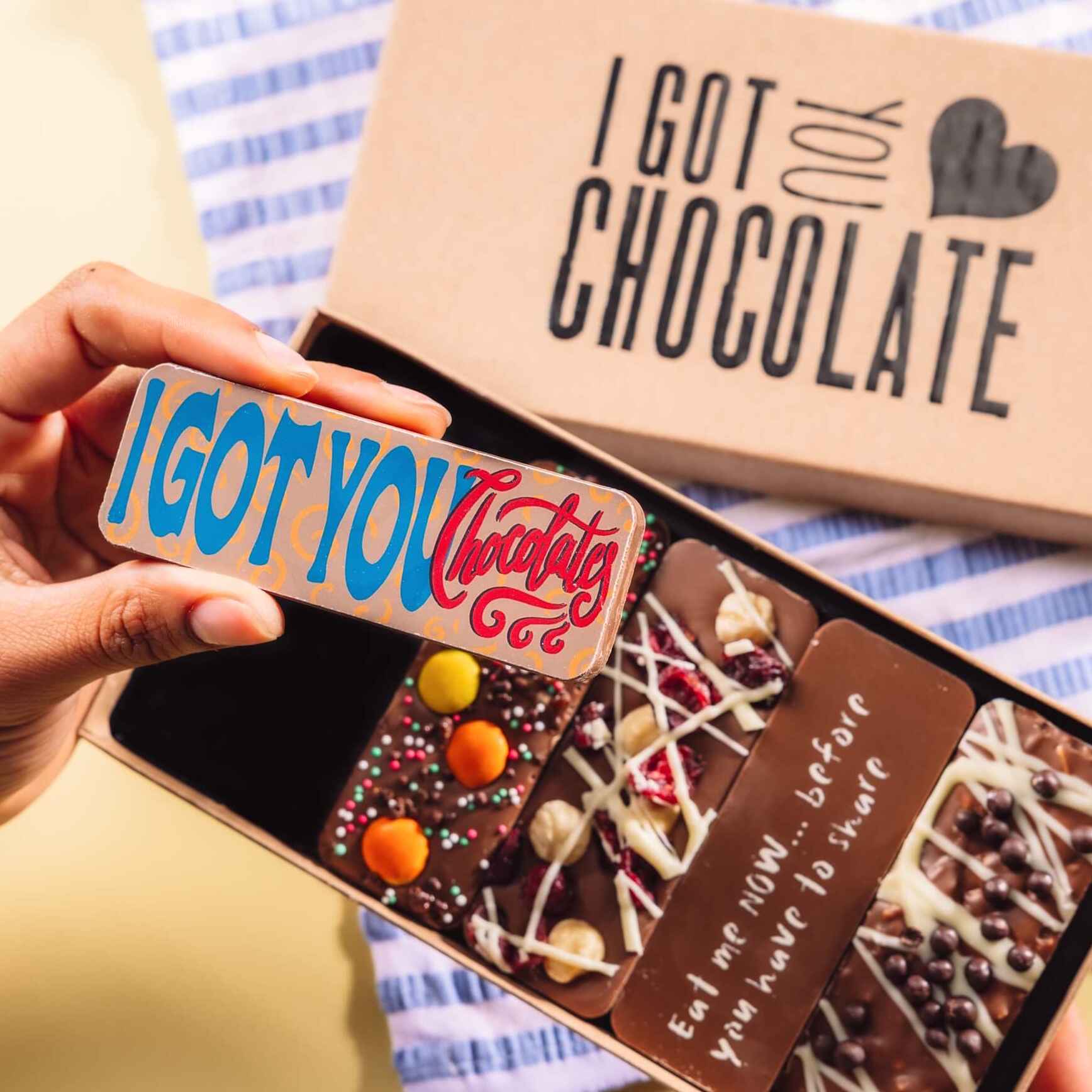 Hand holding up a chocolate bar that says “I Got You Chocolate”, with a gift box of handmade chocolate bars in the background – among the most thoughtful and cheeky I'm sorry gifts