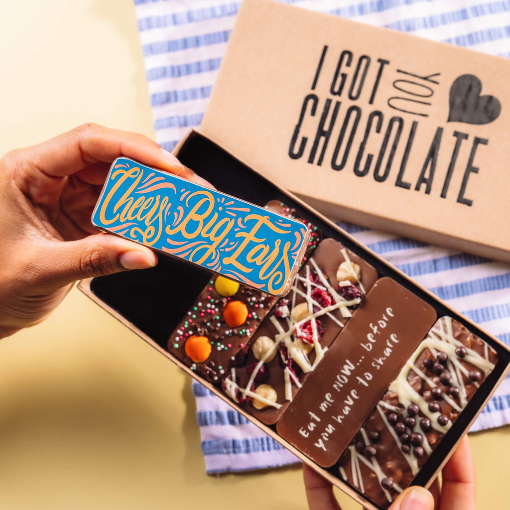 Hand holding a colourful chocolate bar with the message “Cheers Big Ears”, in front of an open gift box of handmade chocolate bars – a fun and thoughtful option for chocolate thank you gifts.