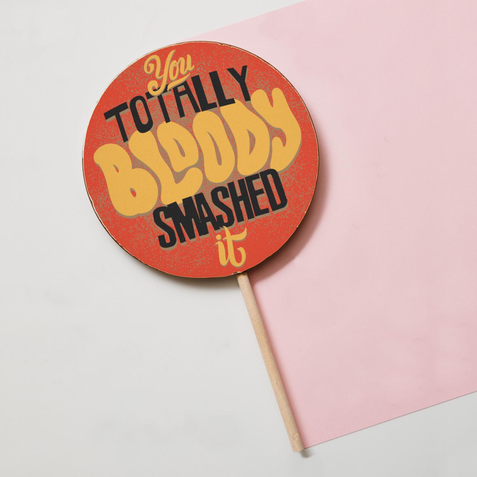 A proud of you gift of a jumbo milk chocolate lollipop with a 'You totally bloody smashed it' message, shown on a pastel background with its wooden stick.