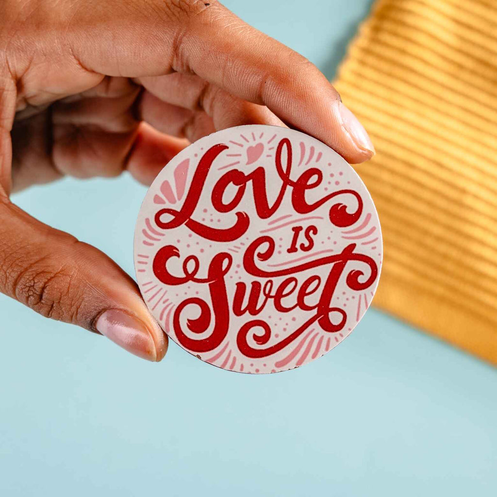 Hand holding a round chocolate with 'Love is Sweet' text on a light blue background