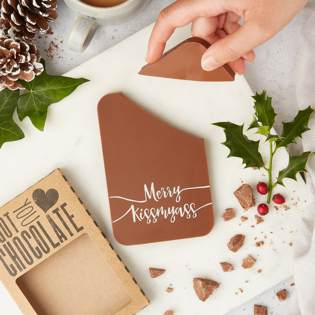Handmade funny Christmas chocolate gift pictured surrounded by holly and pine cones.  The chocolate bar is decorated with a cheeky 'Merry Kissmyass' message.