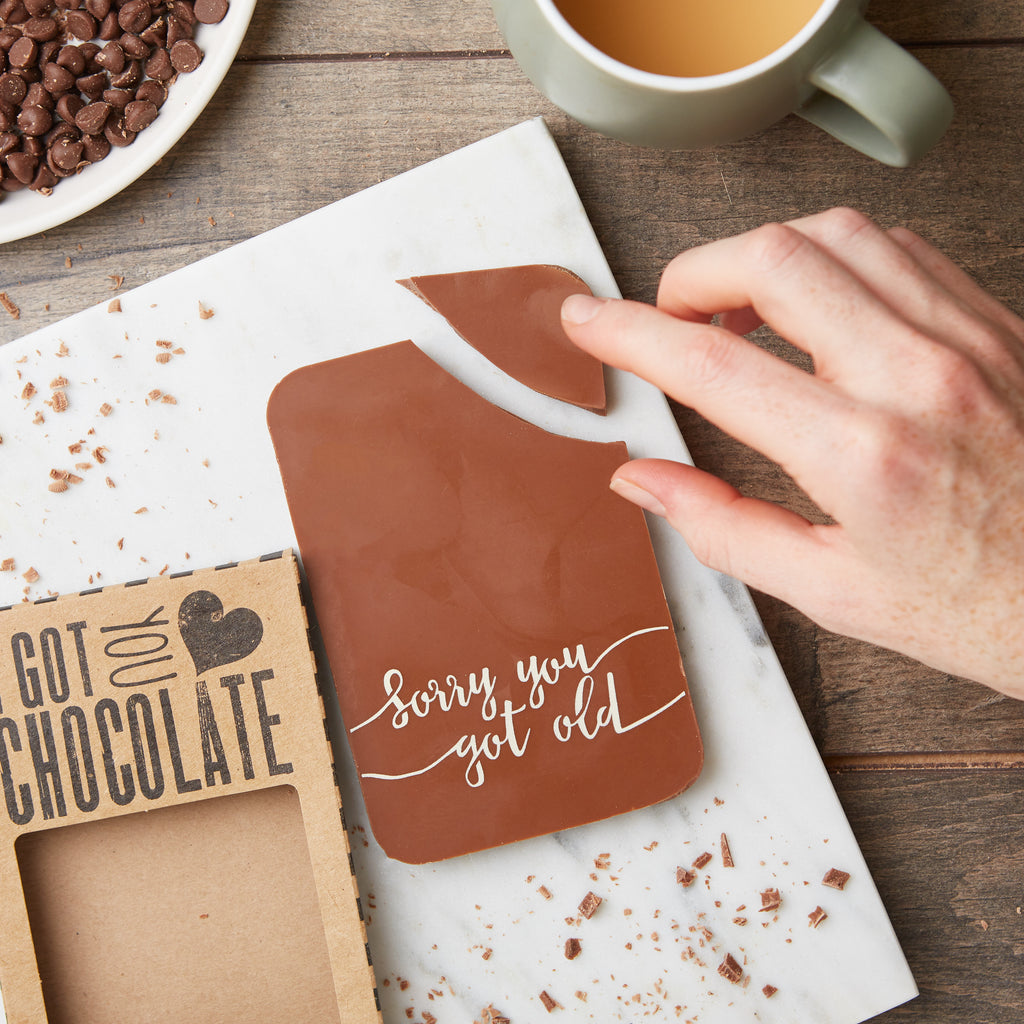 Belgian milk chocolate bar decorated with a 'Sorry you got old' message, shown with a hand stealing a corner away