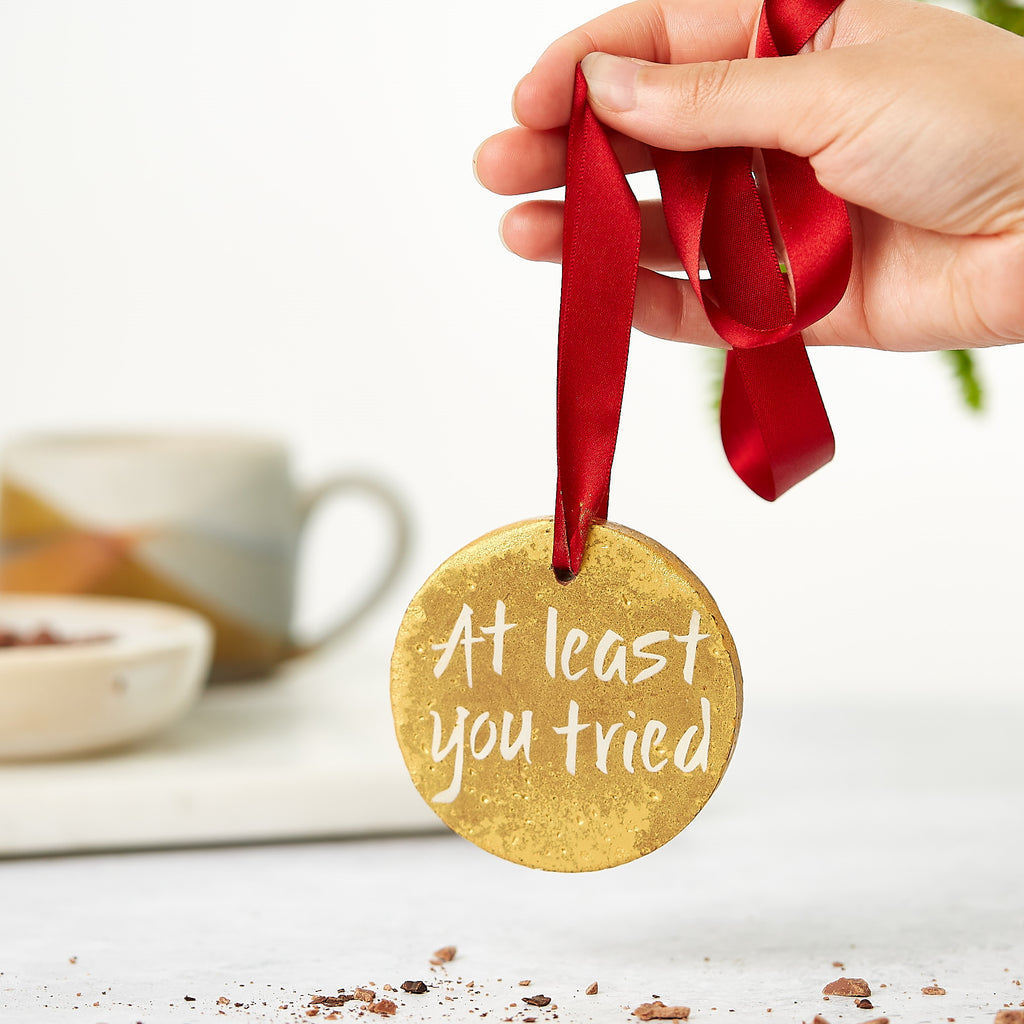 Gold coloured chocolate medal with a cheeky 'At least you tried' message dangling from hand by a red ribbon