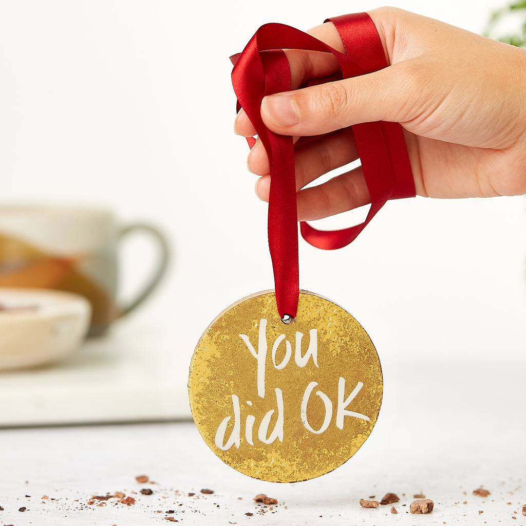 A hand holding the red ribbon of a chocolate gold medal bearing a 'You did OK message